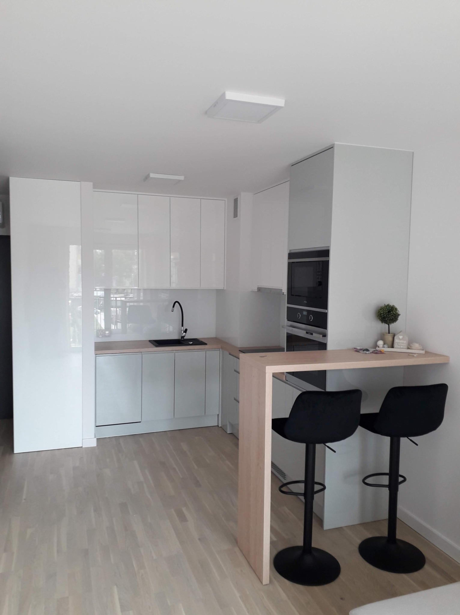 Jasna, nowoczesna kuchnia z białymi, lakierowanymi frontami, czarnym zlewem i drewnianym blatem barowym z dwoma czarnymi hokerami.