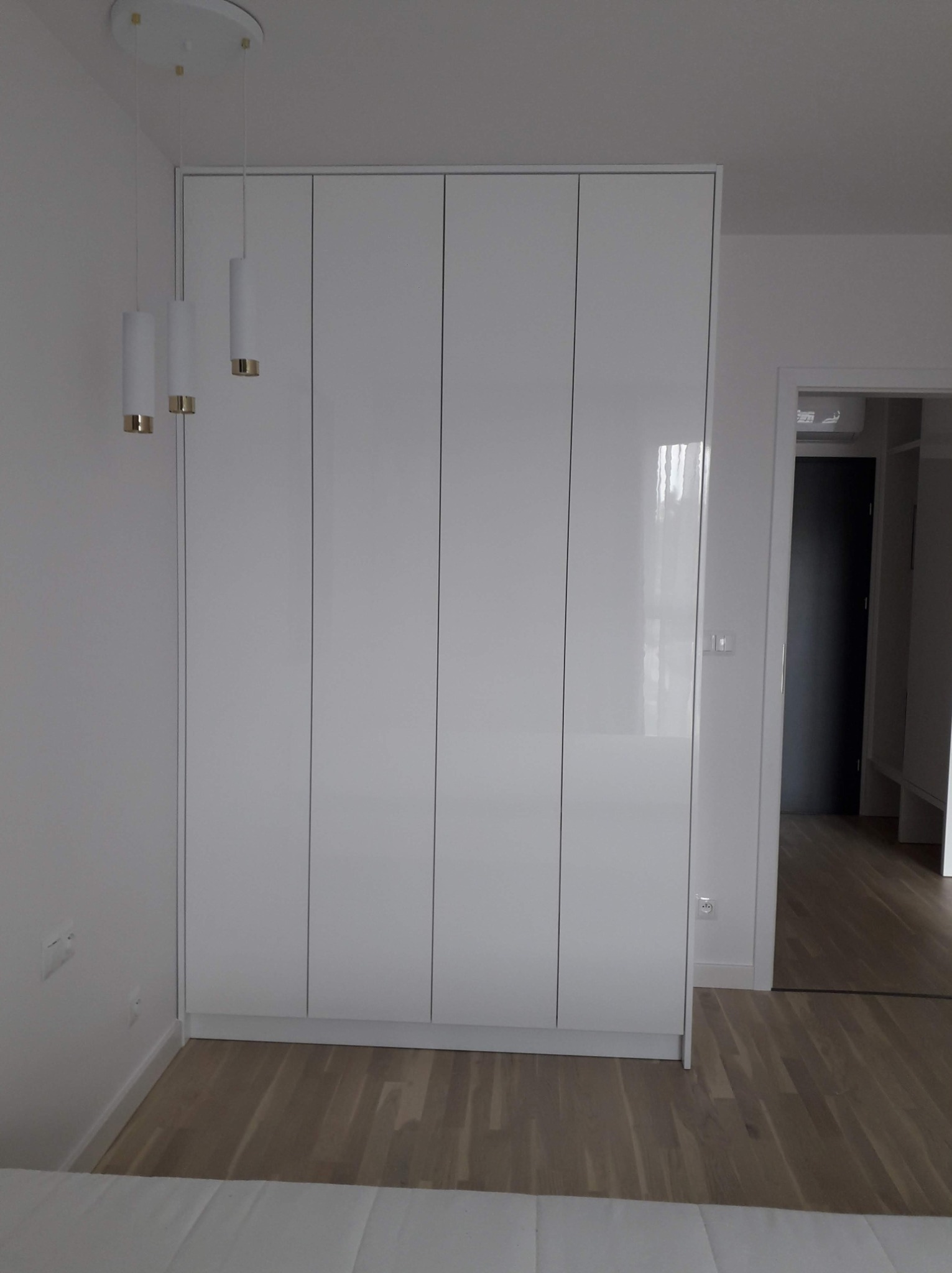 Biała, wysoka szafa z połyskującymi frontami, dopasowana do ściany, z trzema cylindrycznymi lampami wiszącymi z elementami w kolorze złota, widok na wnętrze garderoby w tle.