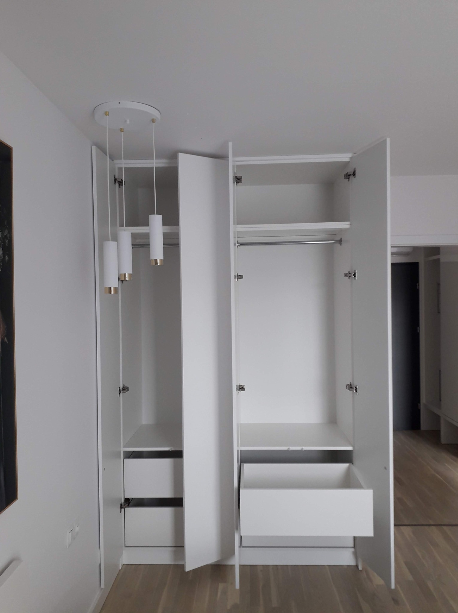 Biała szafa z otwartymi drzwiami, widoczne półki, szuflady i drążek na ubrania, z trzema cylindrycznymi lampami wiszącymi w jednej z komór.