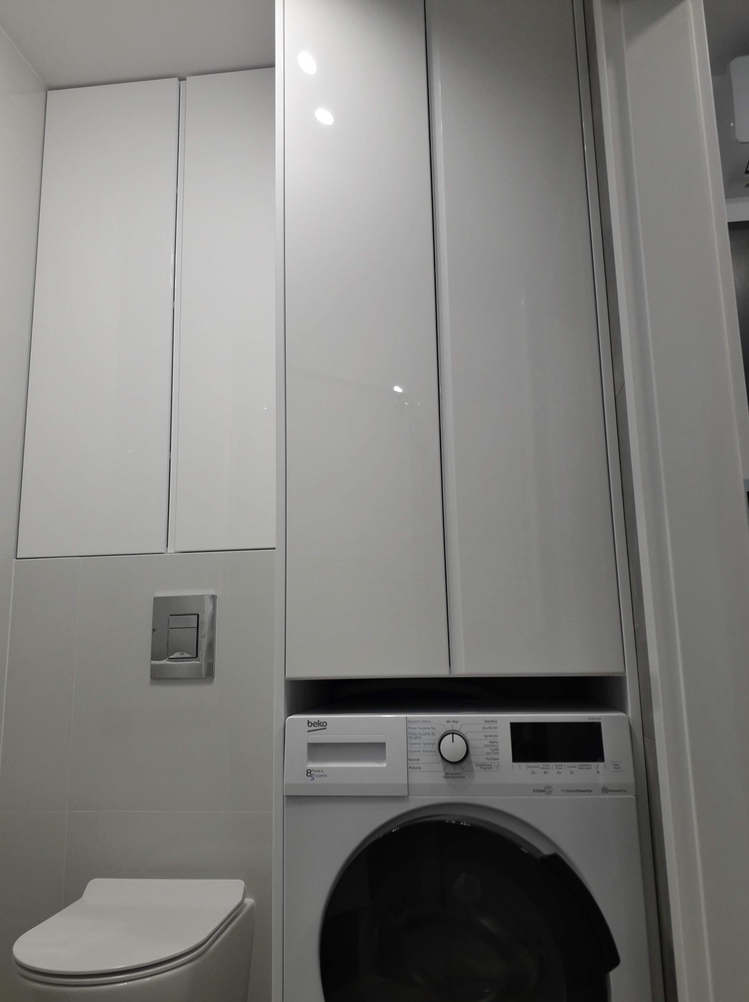 Biała pralka Beko w zabudowie meblowej z połyskującymi białymi frontami, widoczny fragment toalety i przycisku spłuczki, minimalistyczne wnętrze łazienki.