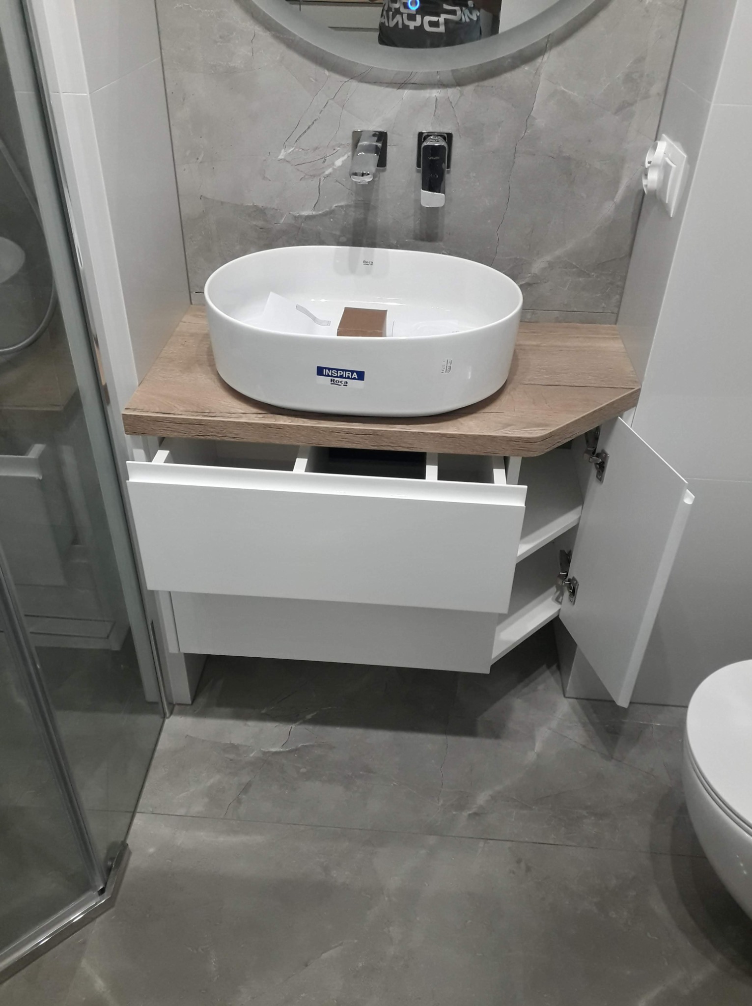 Nowoczesna łazienka z okrągłą białą umywalką nablatową Roca Inspira na szafce z jasnego drewna z otwartą szufladą i uchylnymi drzwiczkami, szare płytki imitujące marmur na ścianie i podłodze.