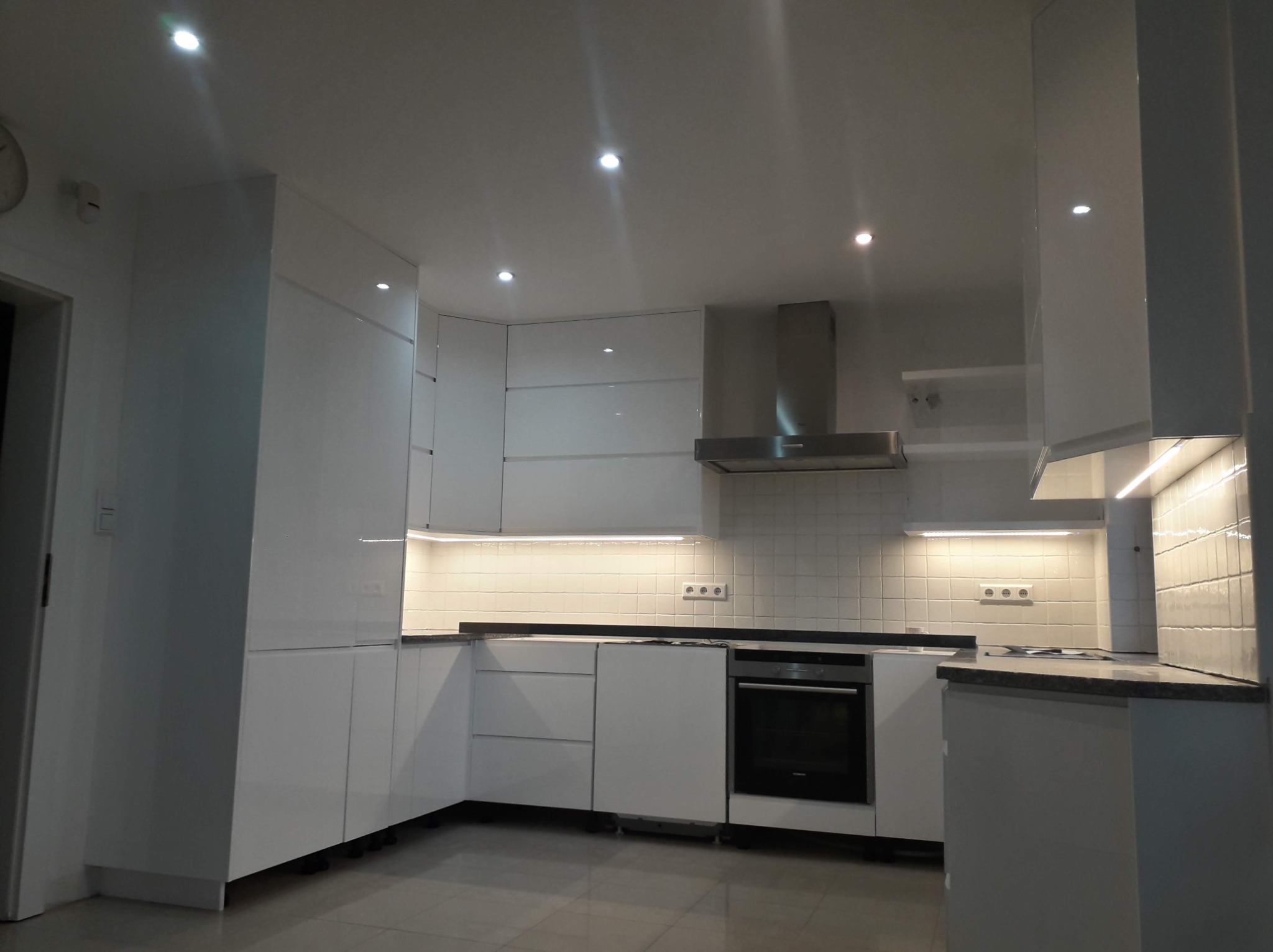 Biała, nowoczesna zabudowa kuchenna z oświetleniem LED podszafkowym i srebrnym okapem, widok na fronty szafek i piekarnik.