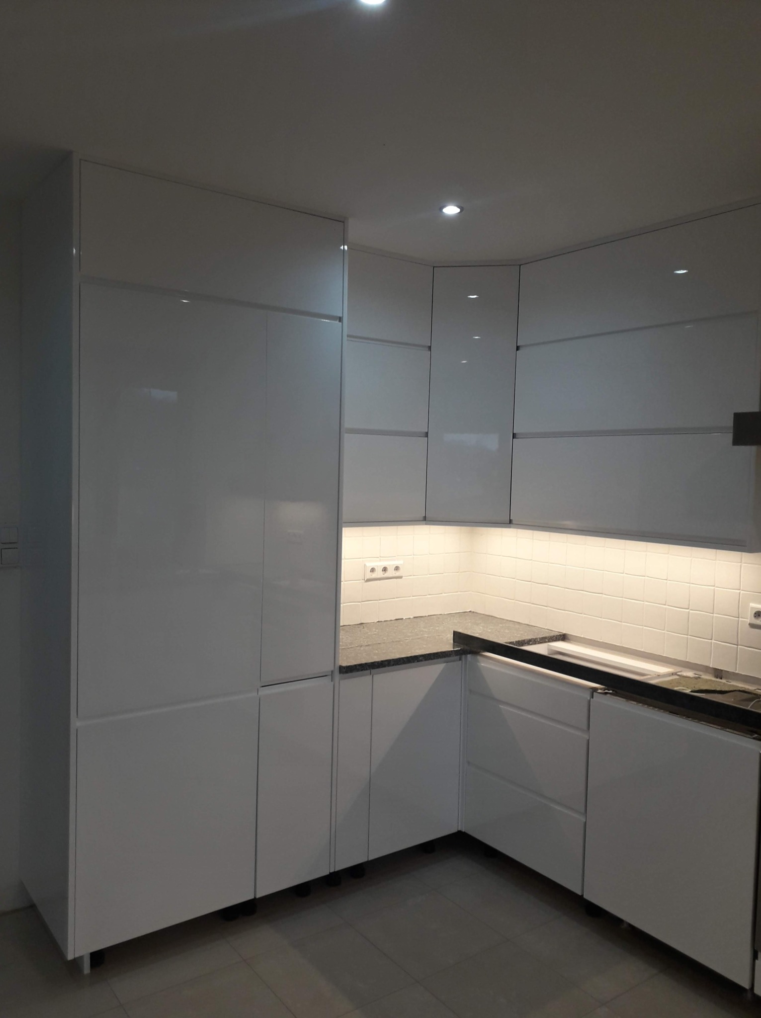 Biała, lakierowana kuchnia na wymiar z czarnym blatem i oświetleniem podszafkowym, widok z góry na narożną zabudowę.