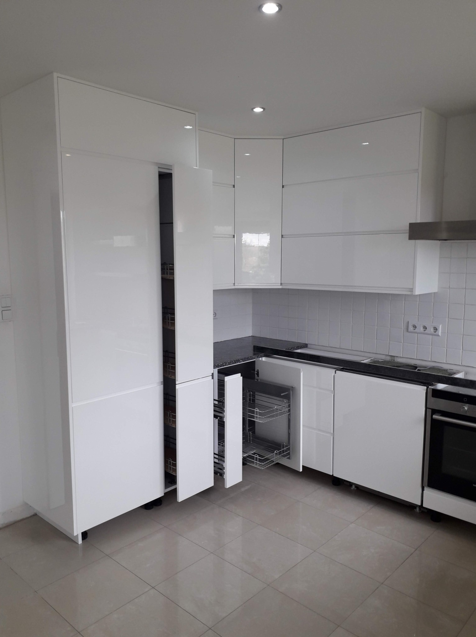 Biała, błyszcząca zabudowa kuchenna z wysuwanymi koszami cargo w wysokiej szafce i pod blatem, na tle białych płytek.