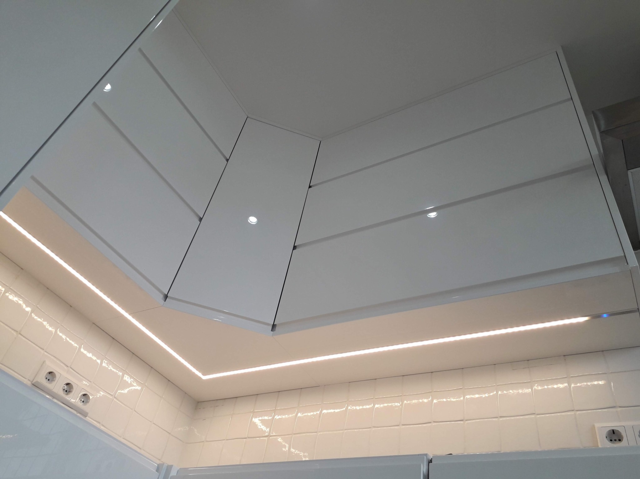 Białe, lakierowane szafki kuchenne podwieszane, oświetlone listwą LED, z widocznymi gniazdkami elektrycznymi na białej, kwadratowej glazurze.