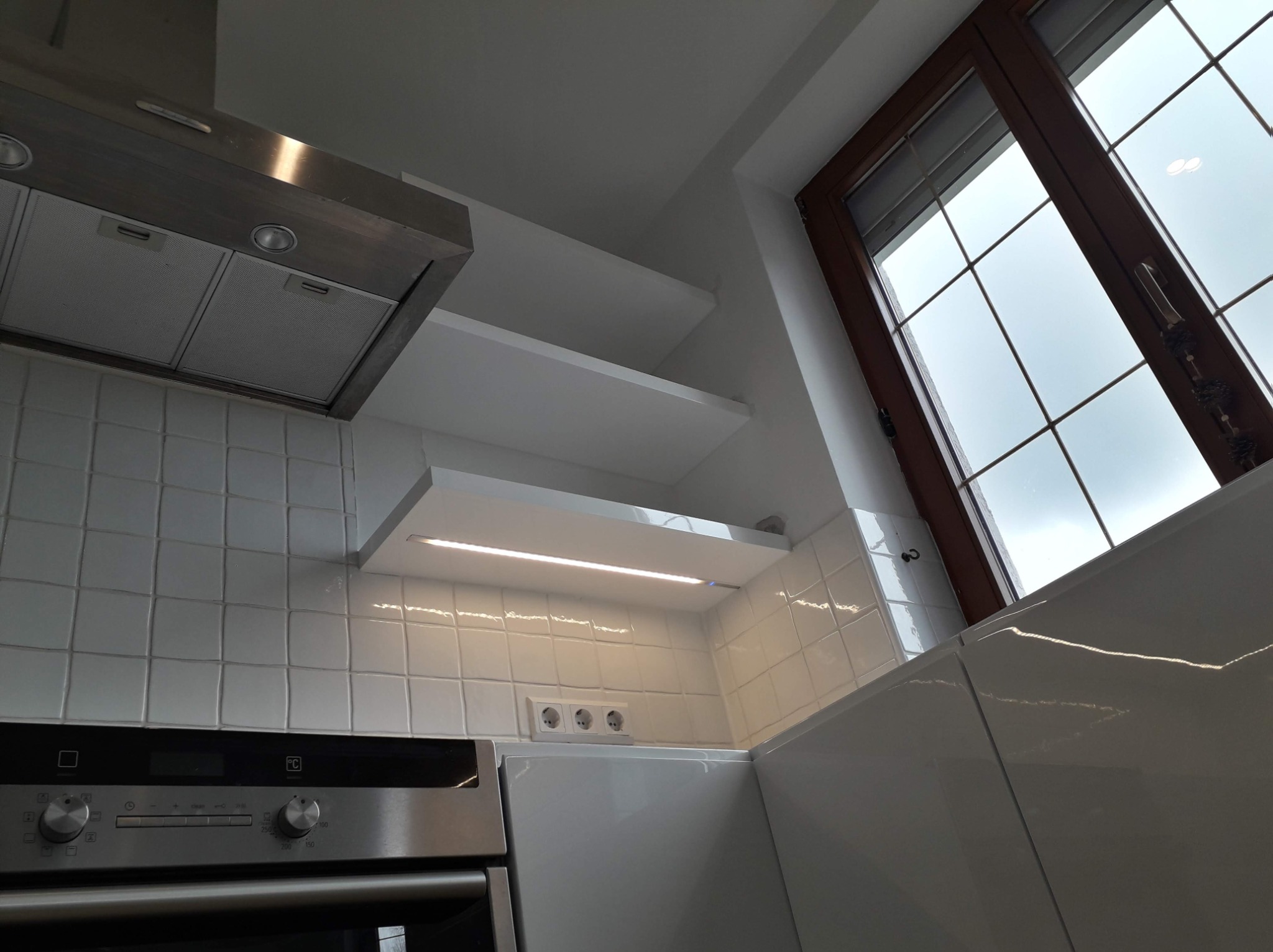 Fragment białej kuchni z okapem, piekarnikiem, białymi półkami z oświetleniem LED i oknem z brązową ramą.