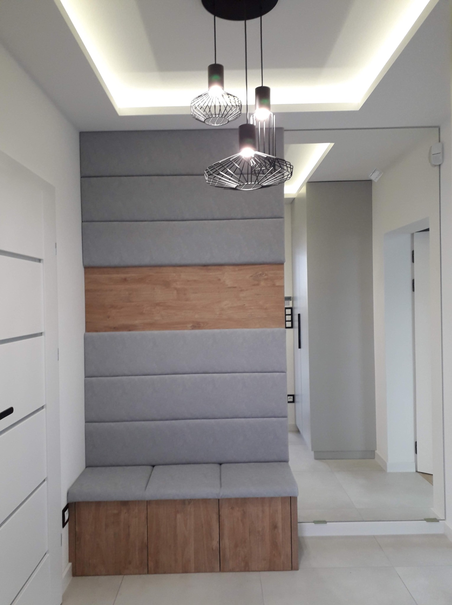 Przedpokój z siedziskiem w zabudowie, tapicerowany panel ścienny z drewnianym akcentem, nowoczesne lampy wiszące i lustro na ścianie.