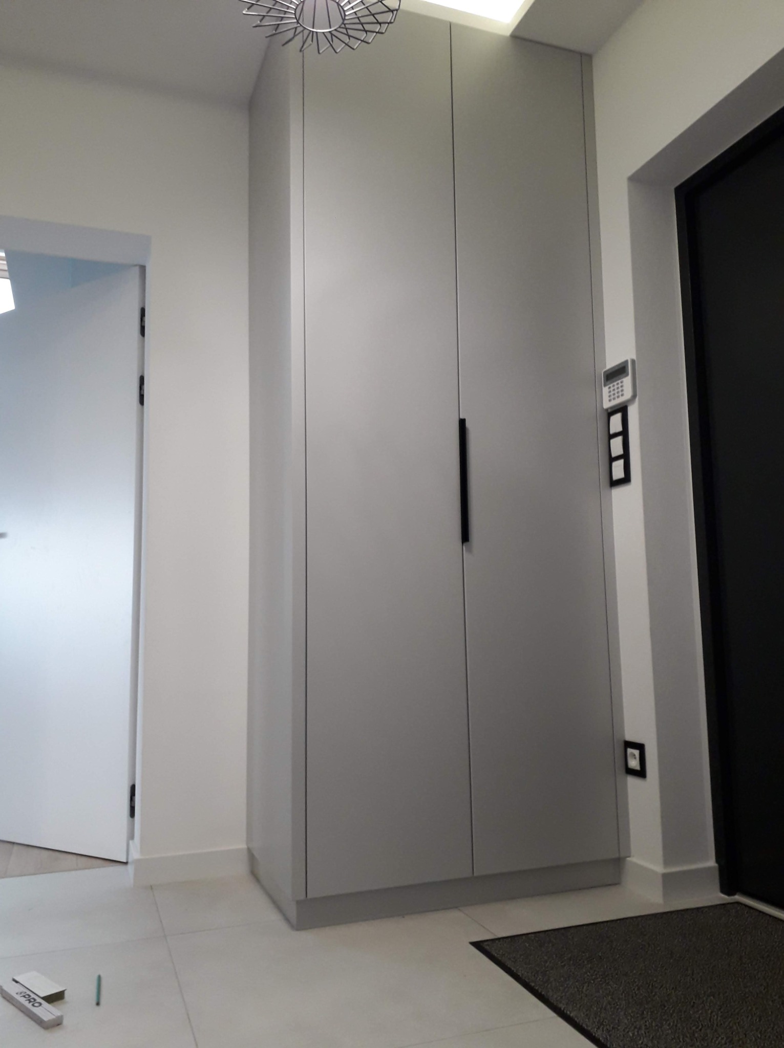 Wysoka, szara szafa wnękowa z długimi, pionowymi uchwytami w przedpokoju z białą podłogą i drzwiami, widoczny panel sterowania alarmem.