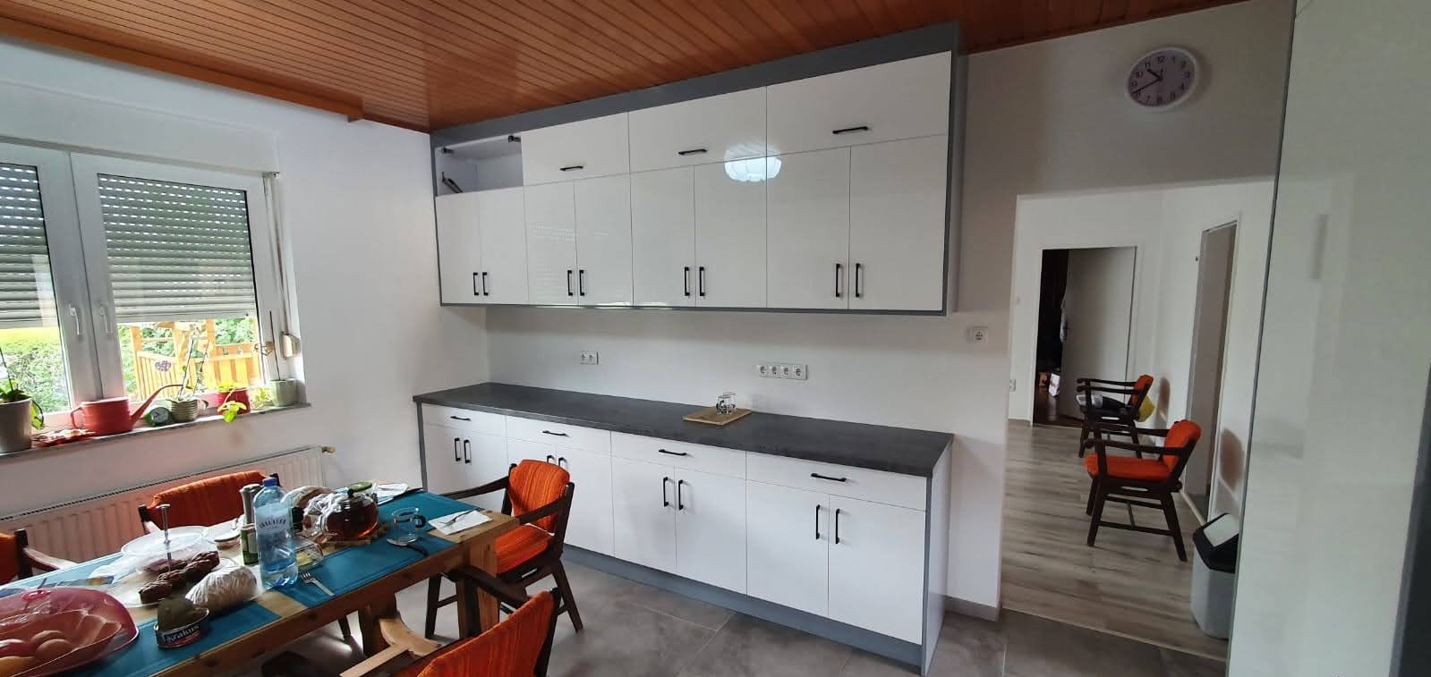 Nowoczesna kuchnia z białymi szafkami z szarymi akcentami i blatem, widoczny stół z posiłkiem, drewniany sufit i zegar na ścianie.