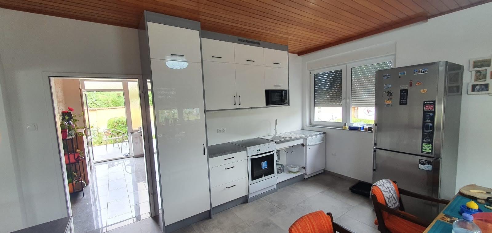 Jasna kuchnia z białymi szafkami z szarymi akcentami, widok na ogród przez otwarte drzwi balkonowe, lodówka z magnesami i zdjęciami.