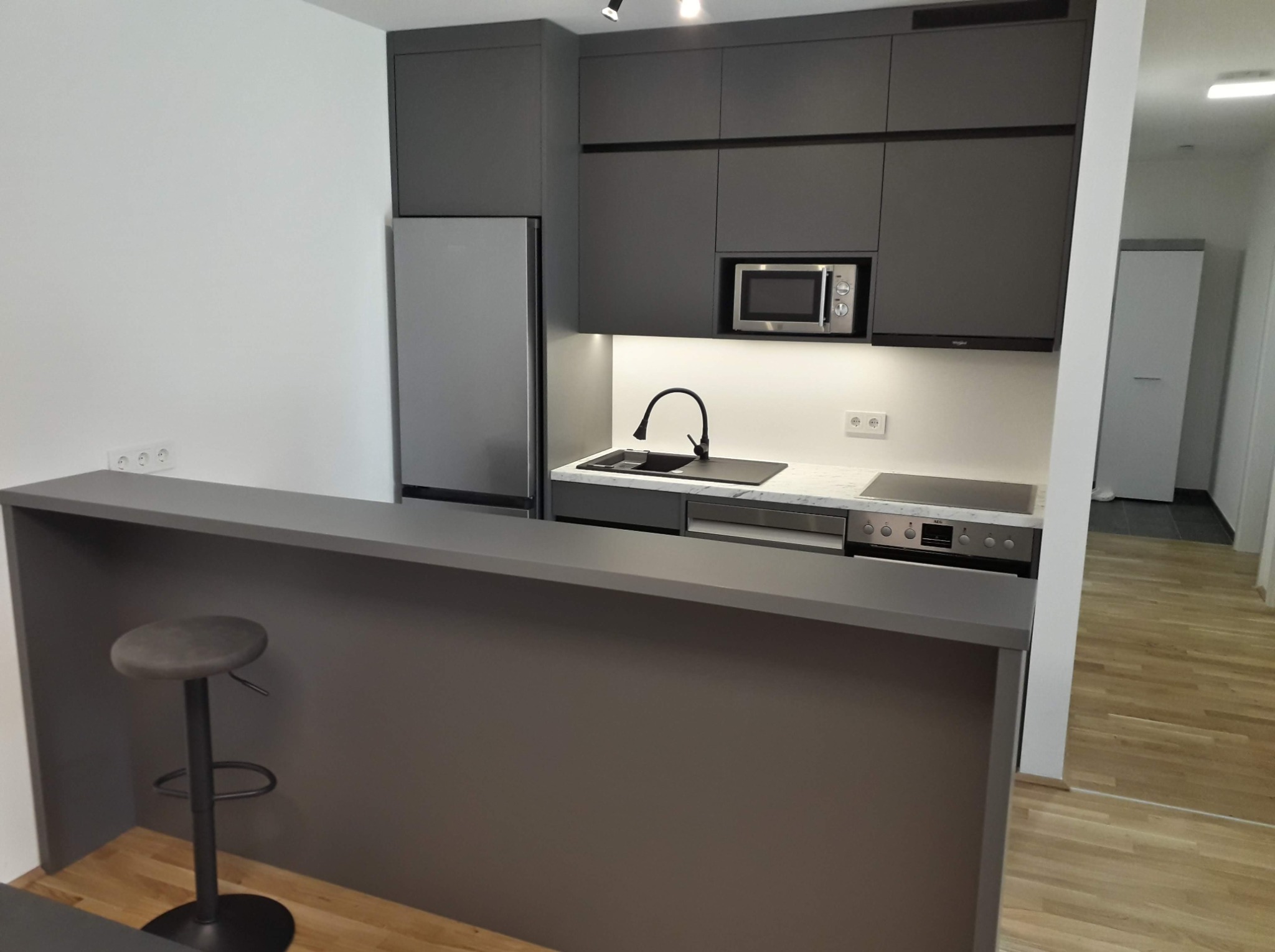 Nowoczesna kuchnia w odcieniach szarości z wyspą i stołkiem barowym, widok na płytę indukcyjną i mikrofalówkę.