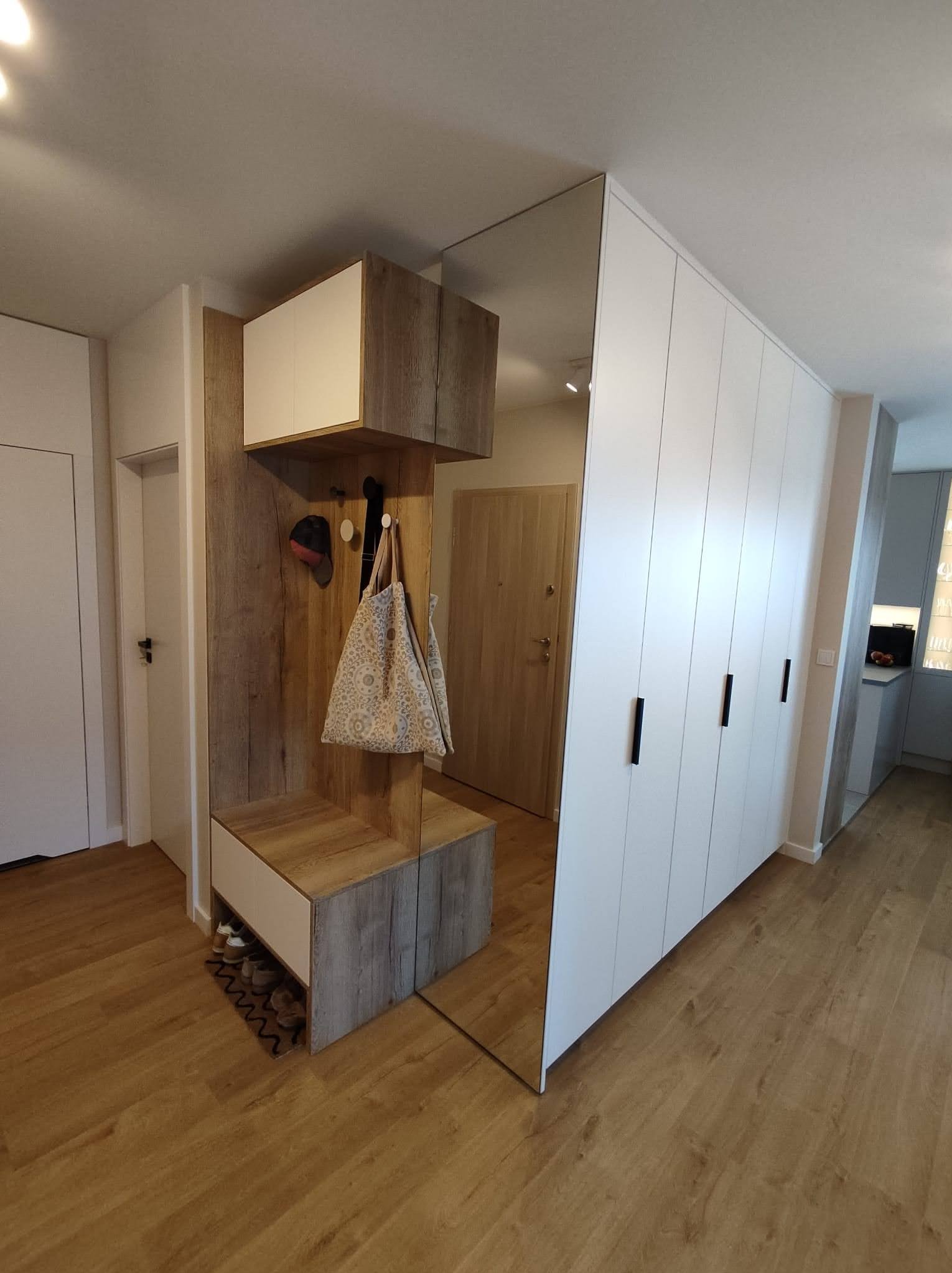Nowoczesny przedpokój z szafą wnękową z lustrem, siedziskiem i półkami w odcieniach bieli i drewna, torba na wieszaku, pod spodem buty.