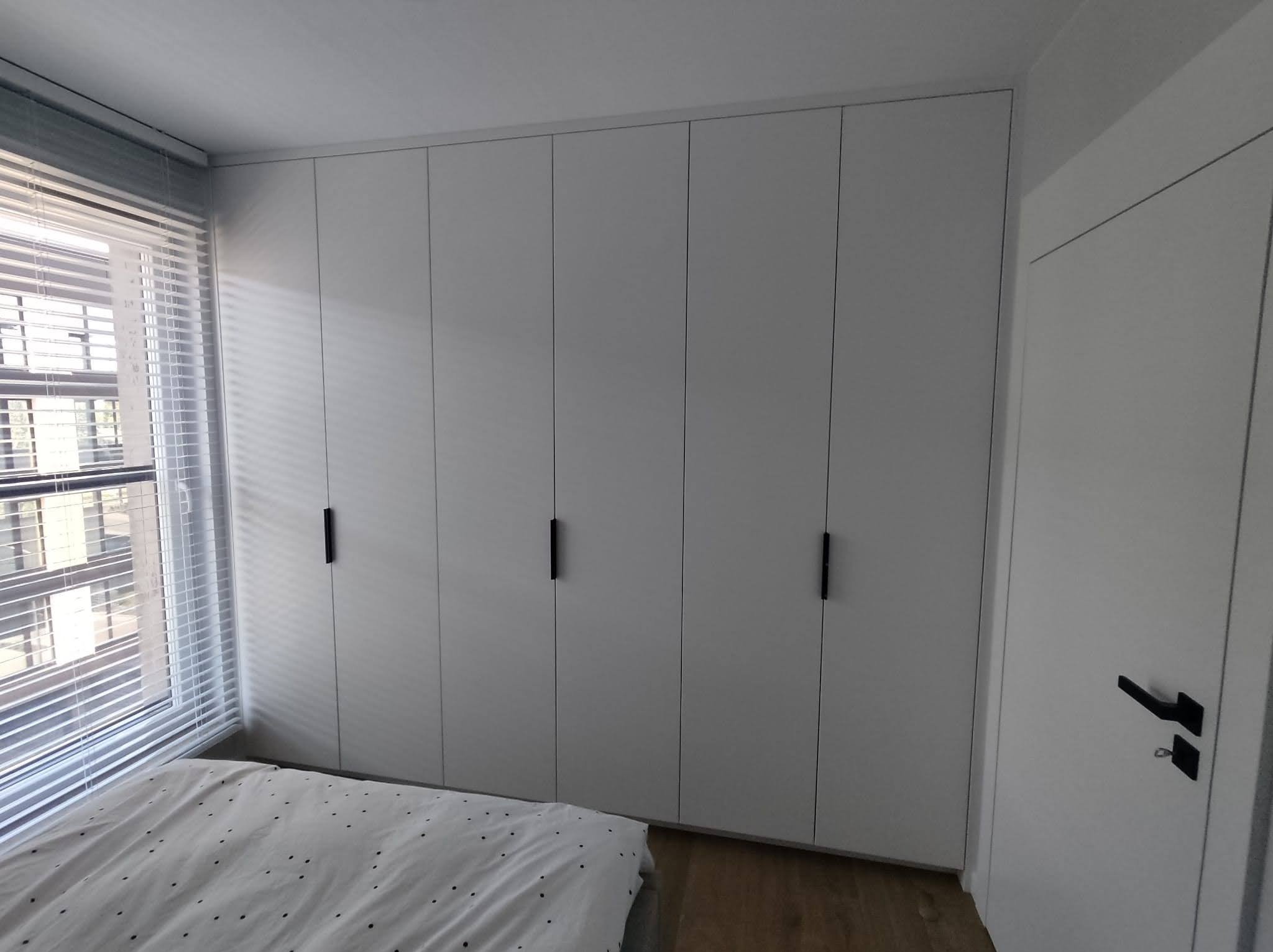 Biała, zabudowana szafa wnękowa z czarnymi, minimalistycznymi uchwytami, widoczna w sypialni z oknem przysłoniętym żaluzjami i fragmentem łóżka z pościelą w kropki.