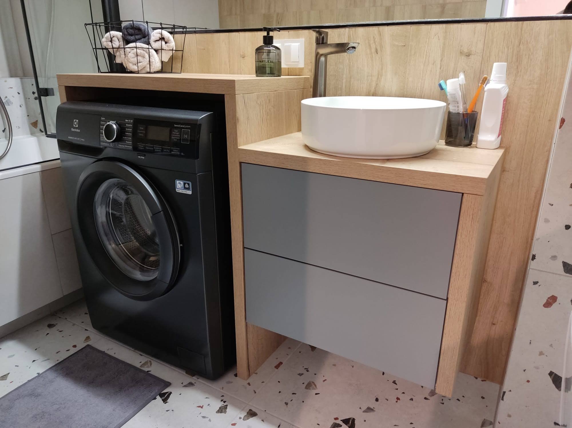 Nowoczesna łazienka z pralką Electrolux obok szafki umywalkowej z szarymi frontami i blatem z jasnego drewna, na podłodze terazzo.