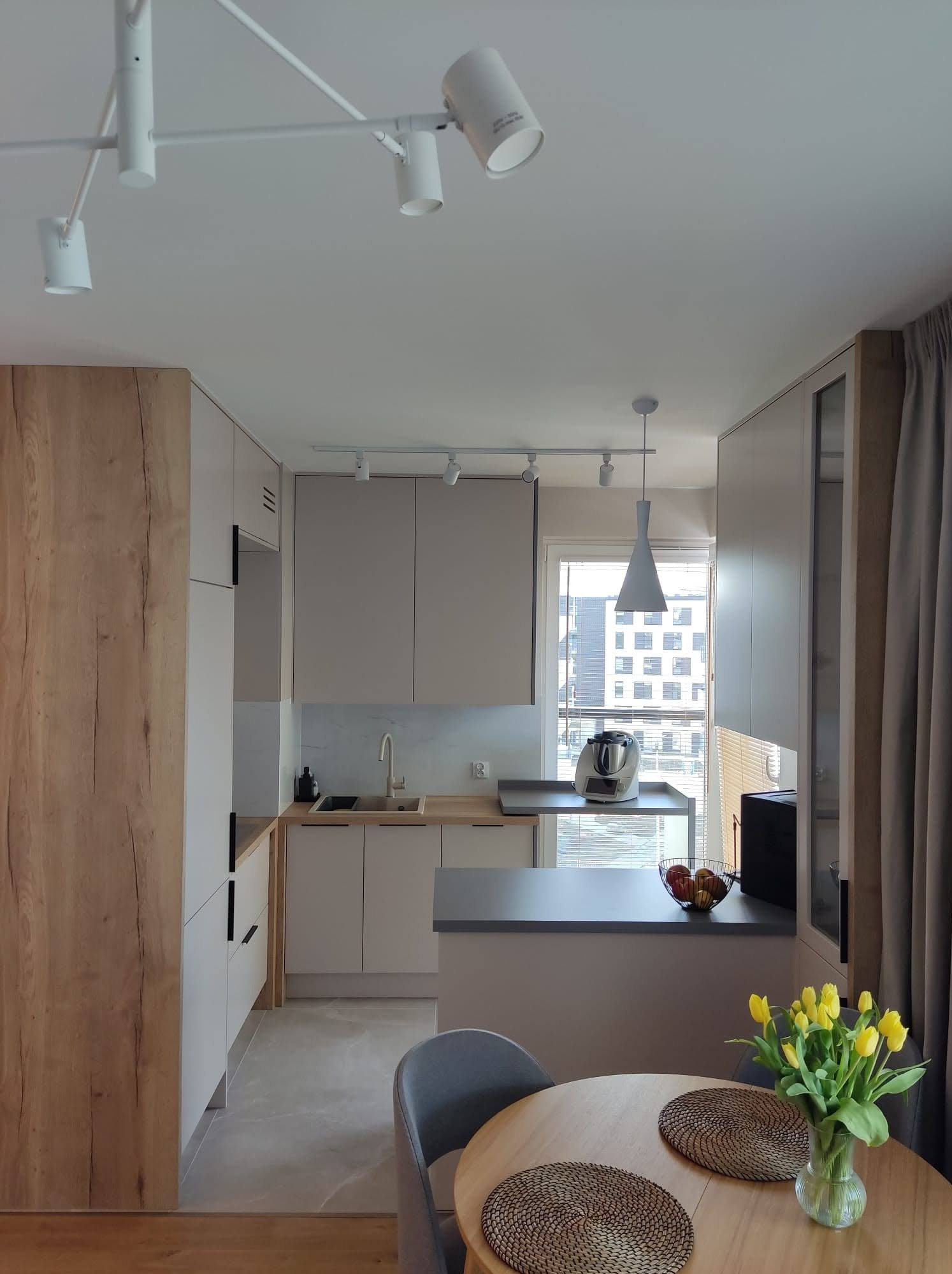 Jasna kuchnia z drewnianymi akcentami, oświetleniem punktowym i wyspą, widok na miasto przez okno, stół z żółtymi tulipanami