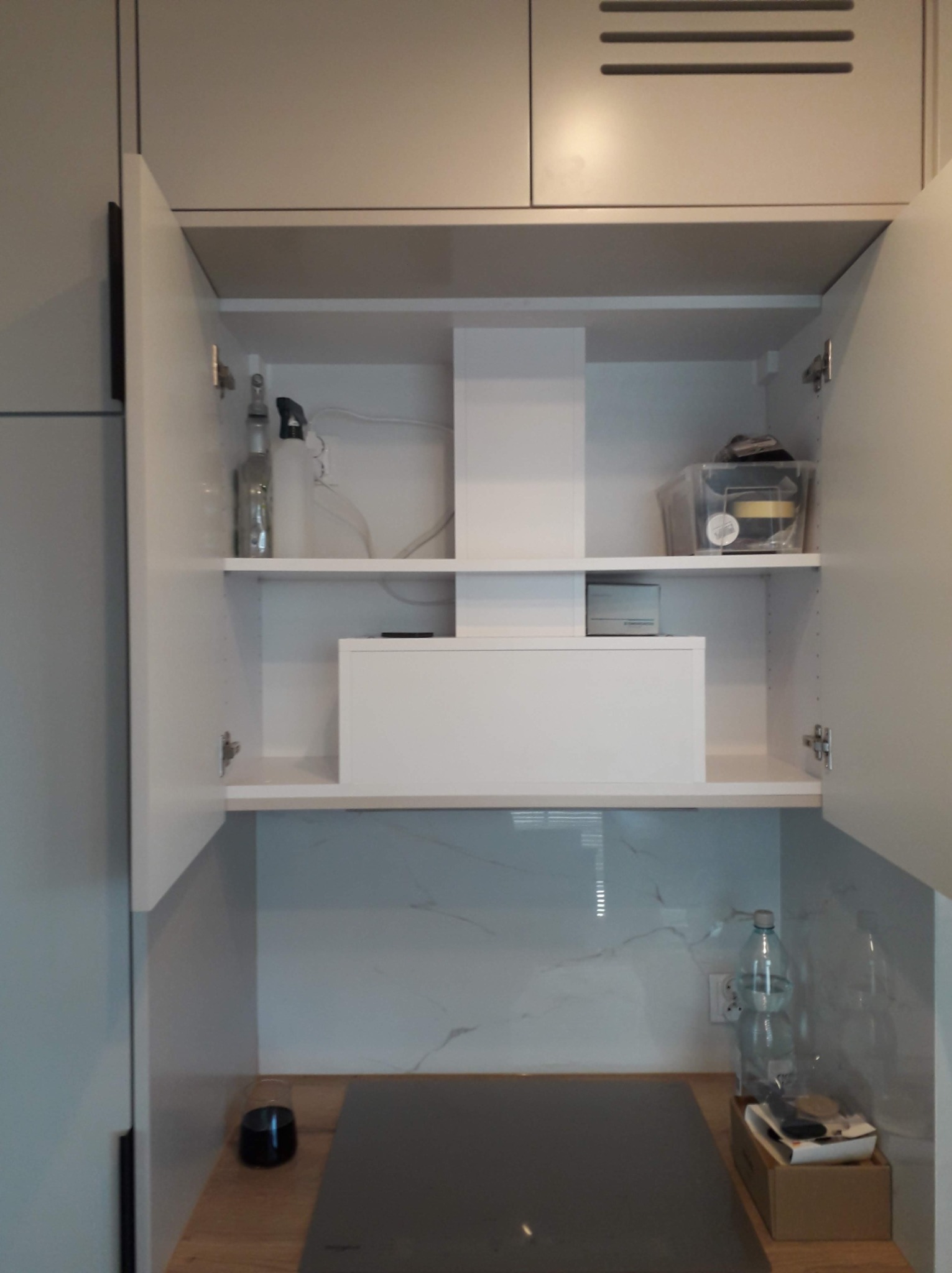 Wnętrze szafki kuchennej z białymi frontami, widoczne półki z różnymi przedmiotami, w tym butelki i pojemniki, nad płytą indukcyjną z blatem imitującym drewno.