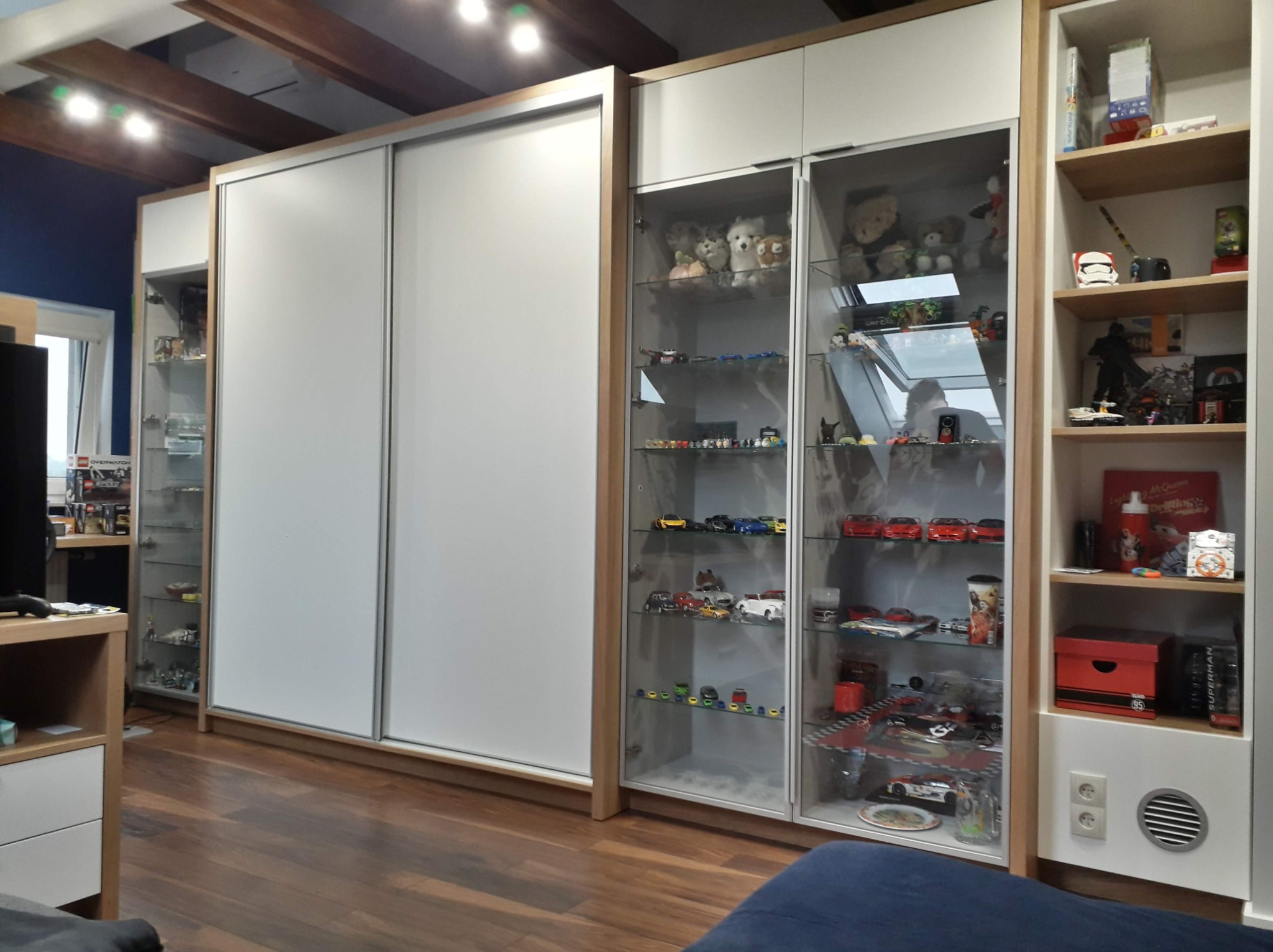 Zestaw mebli: biała szafa przesuwna z jasnym drewnianym obramowaniem, przeszklone witryny z ekspozycją zabawek, pluszaków i modeli samochodów, oraz regał z półkami na zabawki i dekoracje.