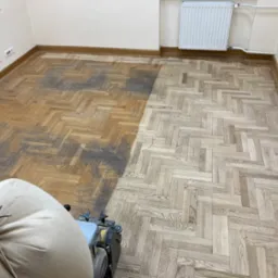 Częściowo odnowiony parkiet w jodełkę, widoczna różnica w kolorze przed i po obróbce. W rogu maszyna do cyklinowania z workiem na pył.