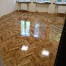 Błyszcząca, odnowiona podłoga z parkietu ułożonego w jodełkę, z widocznymi refleksami światła w przestronnym pokoju z trzema oknami i białymi grzejnikami.