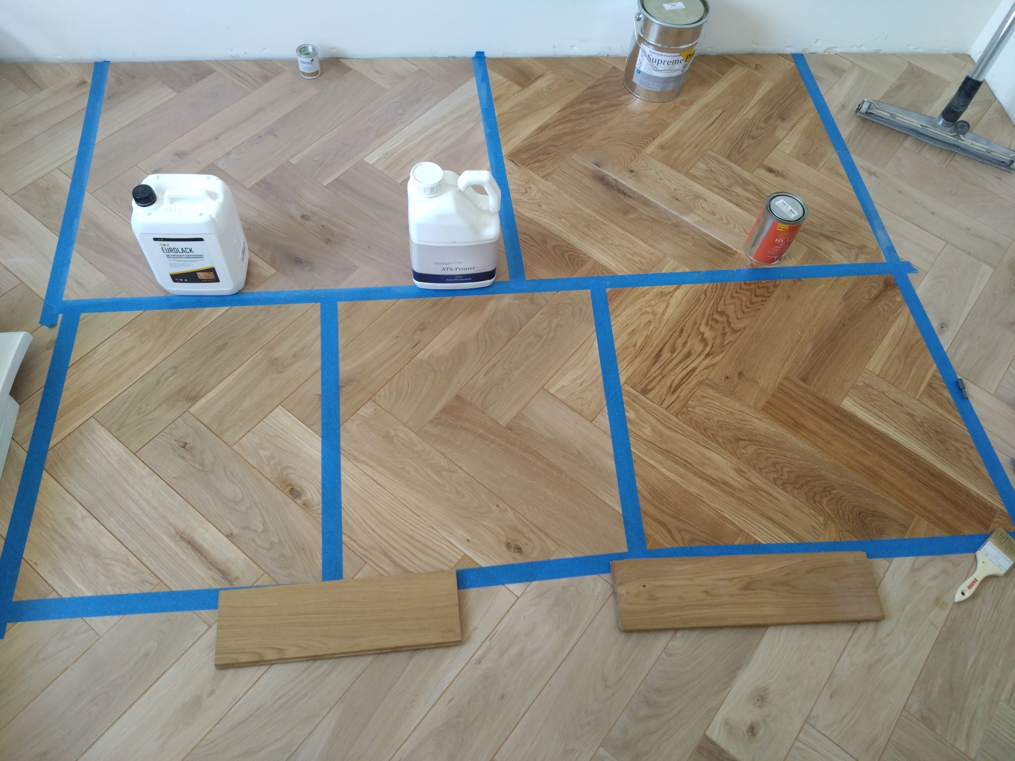 Fragment parkietu ułożonego w jodełkę, podzielony niebieską taśmą malarską na kwadraty, z różnymi próbkami lakierów i preparatów do drewna, widoczne puszki z lakierem, gruntem i szczotka