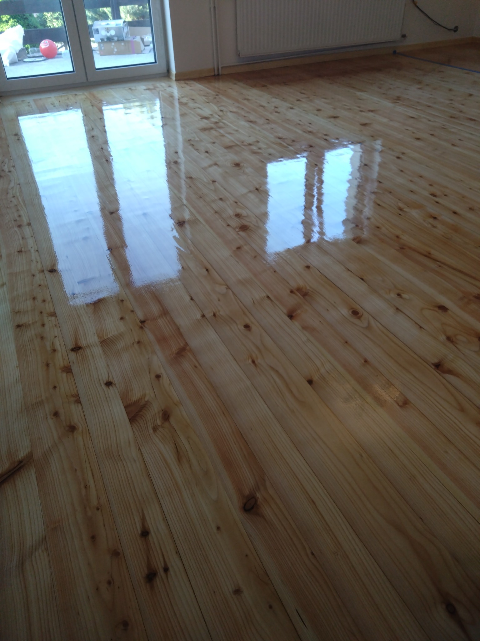 Świeżo polakierowana, jasna podłoga z desek z widocznym odbiciem okna i drzwi balkonowych, ukazująca gładką i lśniącą powierzchnię.