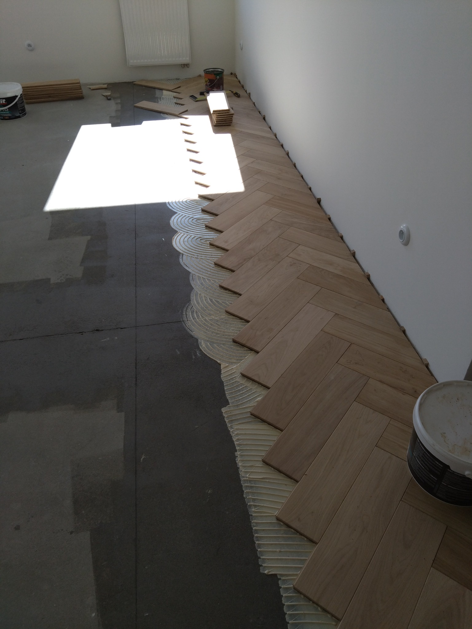 Układanie parkietu w jodełkę: widoczny fragment podłogi z ułożonymi deskami, klejem i niezabezpieczoną, surową posadzką w jasnym wnętrzu.