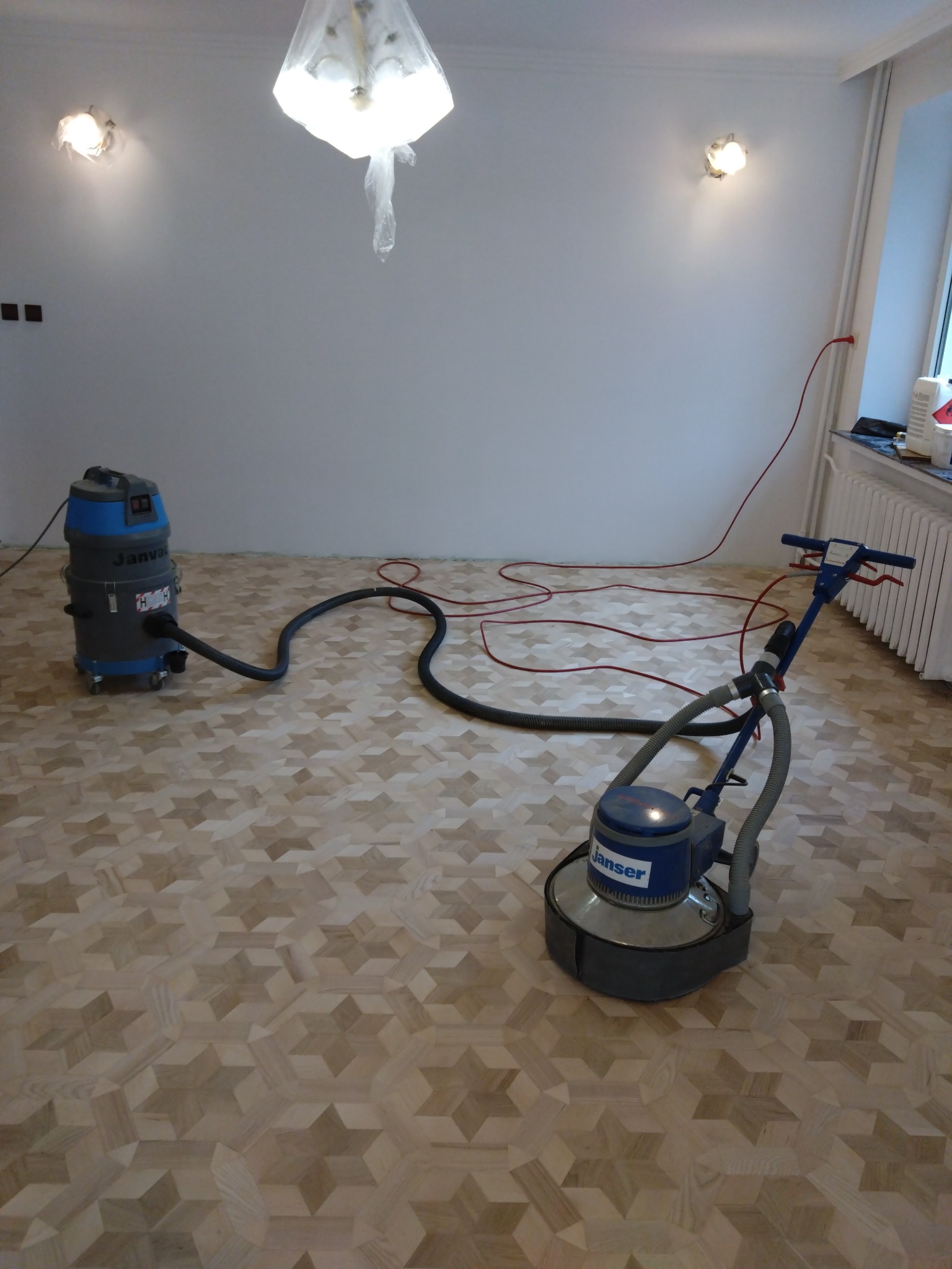 Pokój z parkietem w geometryczny wzór gwiazd, maszyna do szlifowania podłóg Janvac podłączona do odkurzacza przemysłowego, lampa zabezpieczona folią malarską.