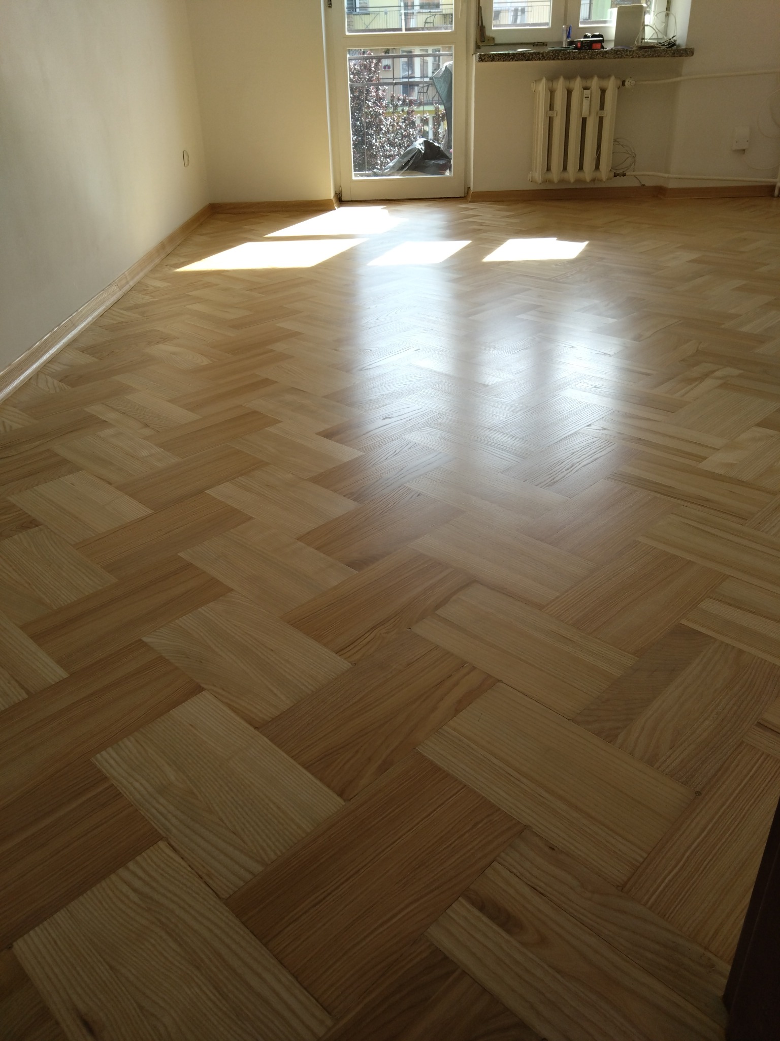 Jasna, nowo ułożona podłoga z parkietu w jodełkę, z widocznymi promieniami słońca wpadającymi przez okno balkonowe, oświetlającymi gładką powierzchnię.