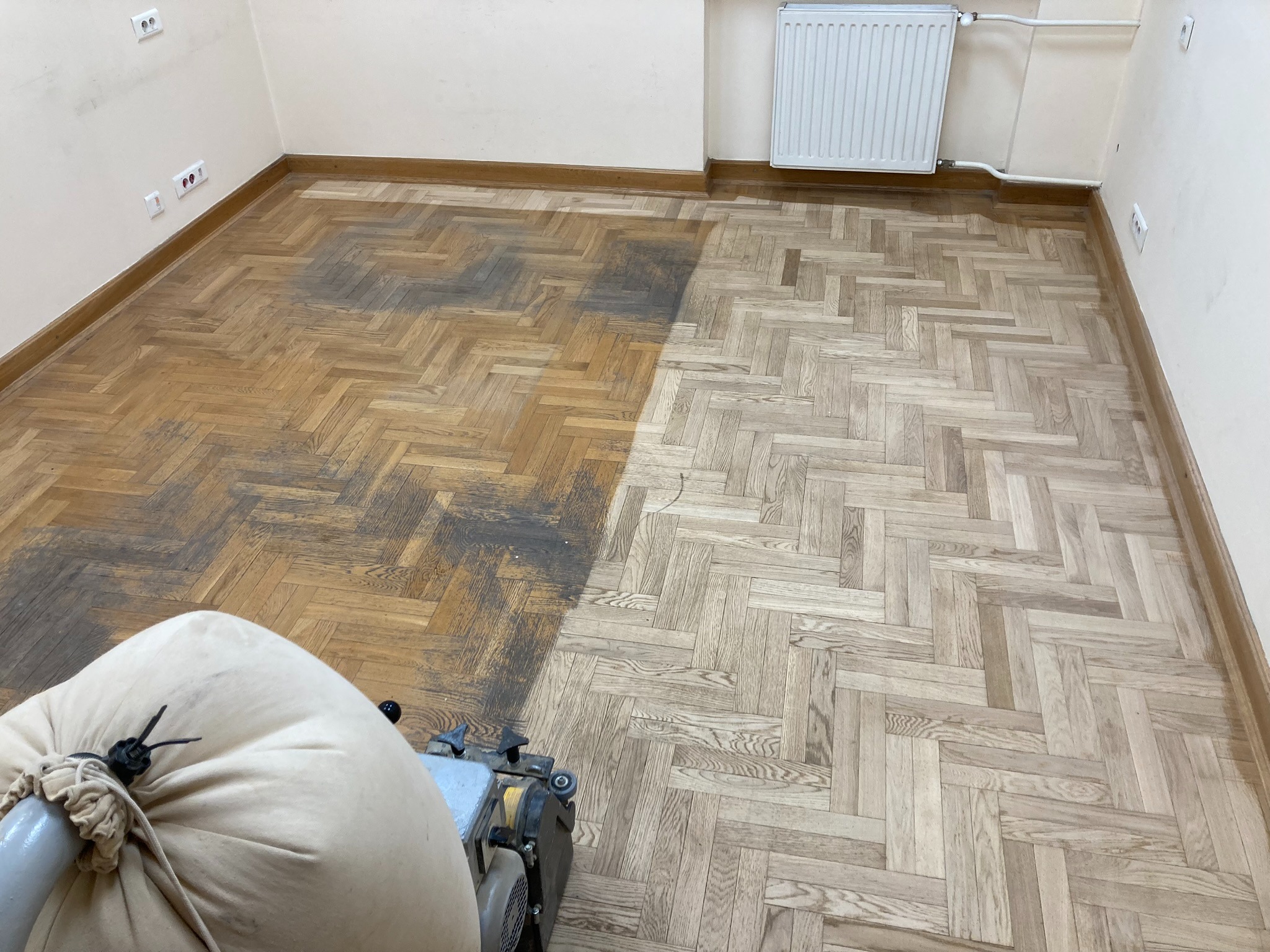 Częściowo odnowiony parkiet w jodełkę, widoczna różnica w kolorze przed i po obróbce. W rogu maszyna do cyklinowania z workiem na pył.