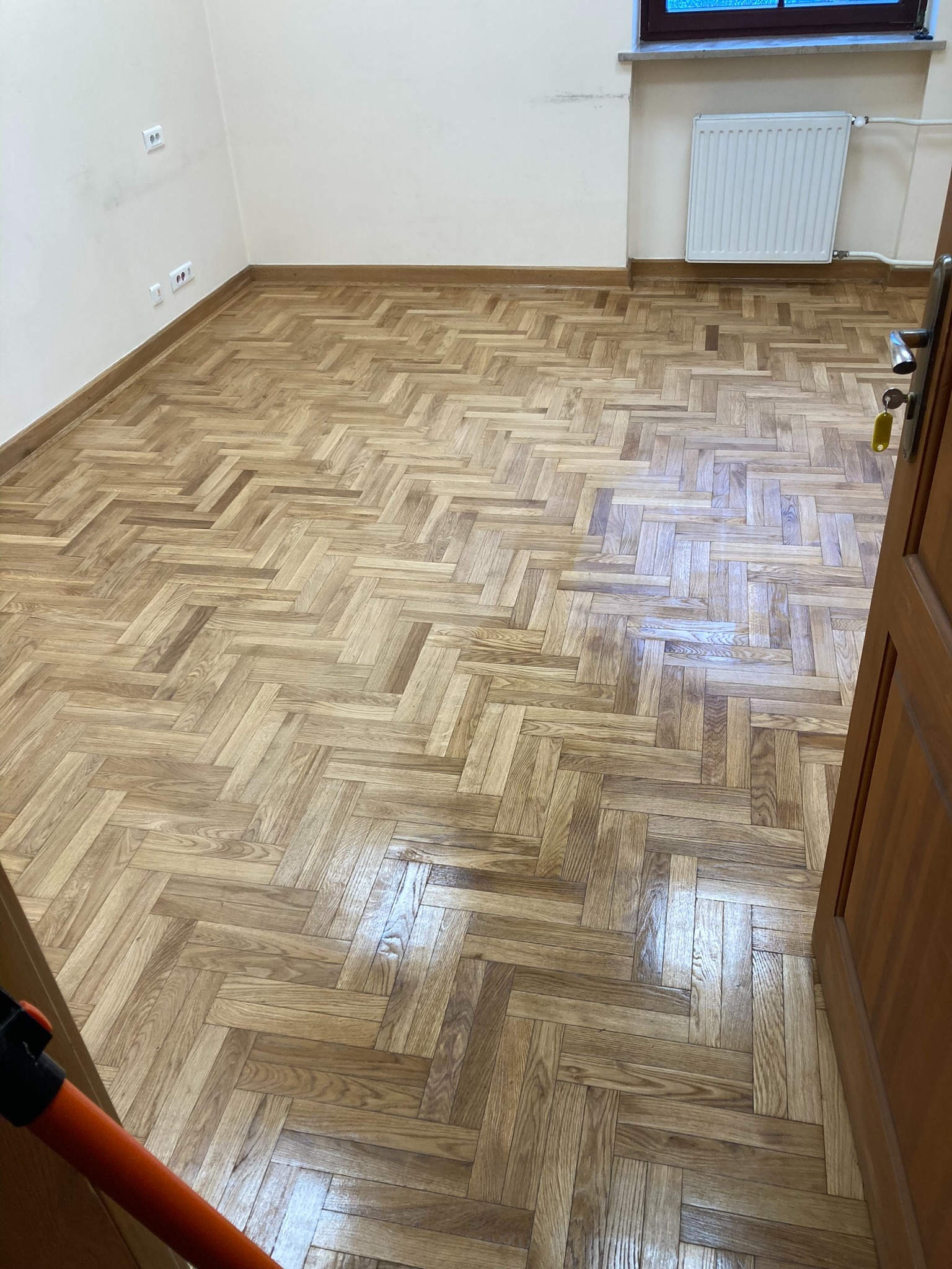 Parkiet ułożony w jodełkę w jasnym pokoju z grzejnikiem. Widoczne gniazdka elektryczne i fragment otwartych drzwi. Podłoga po renowacji, z widocznym połyskiem.