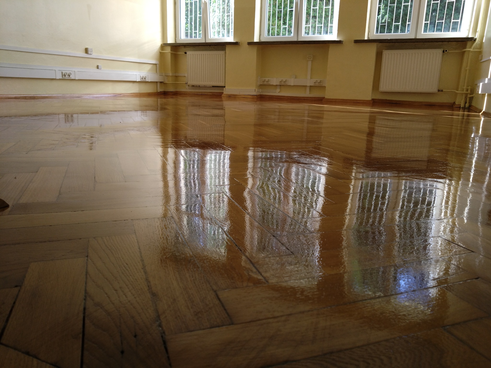 Błyszczący parkiet ułożony w jodełkę, odbijający światło z okien. Widoczne grzejniki pod oknami w jasnym pomieszczeniu. Podłoga po renowacji.