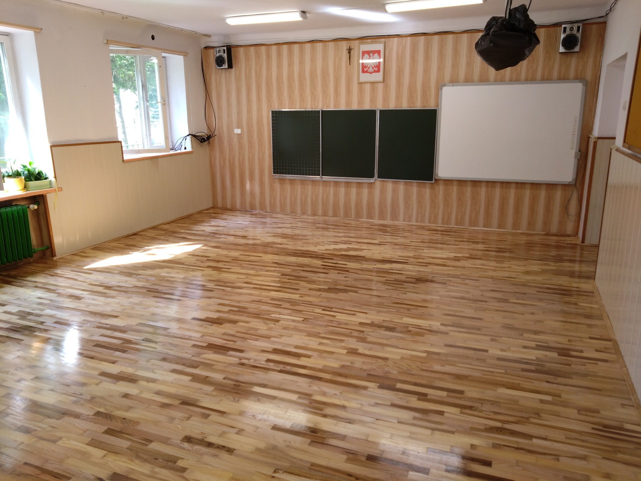 Jasna, drewniana podłoga parkietowa w sali lekcyjnej z zielonym grzejnikiem i tablicami. Widoczne refleksy światła na powierzchni podłogi.