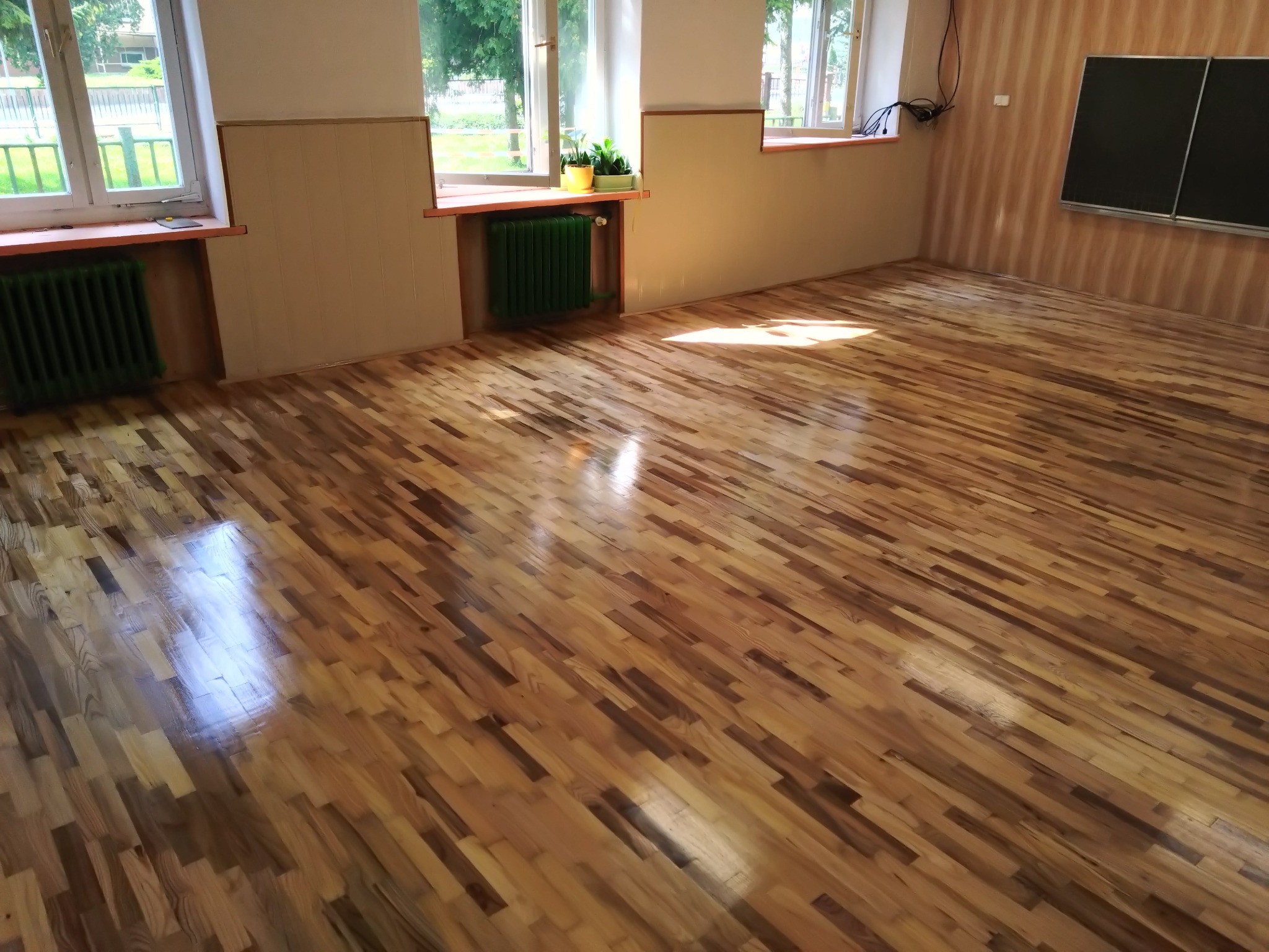 Błyszcząca, odnowiona podłoga parkietowa w jasnym pomieszczeniu z oknami i zielonymi grzejnikami. Widoczne refleksy światła na parkiecie.