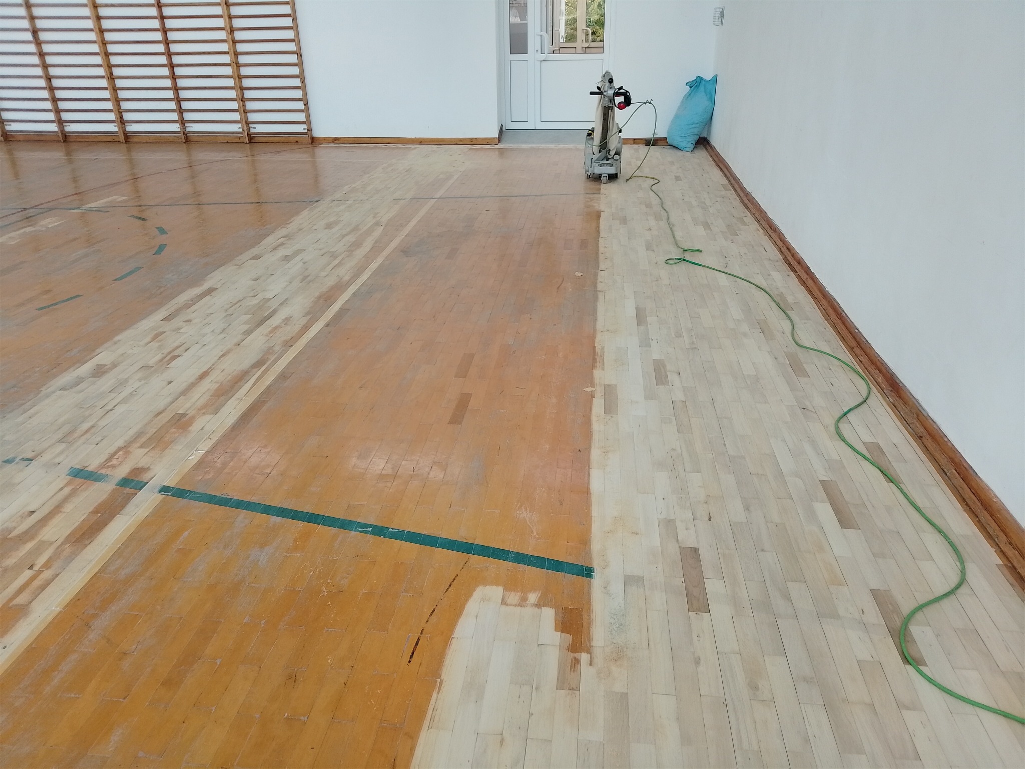 Sala gimnastyczna z częściowo odnowioną drewnianą podłogą. Widoczna maszyna do cyklinowania i fragment ściany z drabinkami. Podłoga z zielonymi liniami boiska.