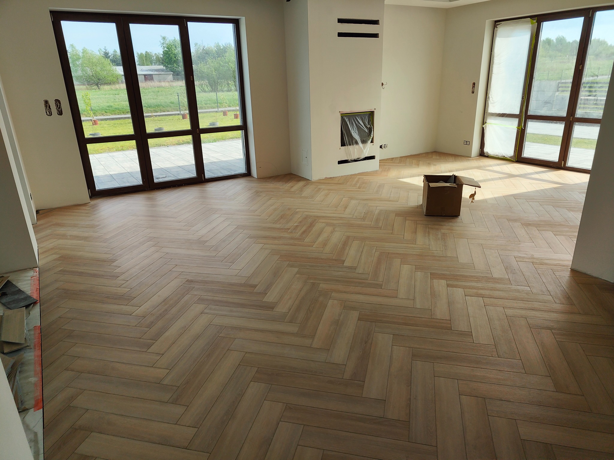 Jasne, drewnopodobne panele ułożone w jodełkę na podłodze przestronnego, jasnego pokoju z dużymi oknami. Widoczny fragment niezakończonej pracy przy ścianie i karton na podłodze.