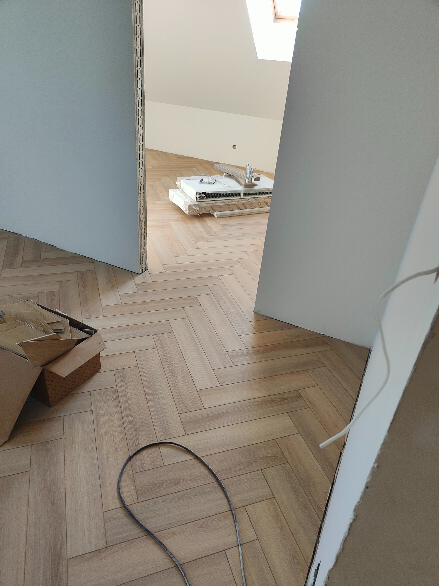 Ukończona podłoga z paneli ułożonych w jodełkę w Sędziszowie Małopolskim. Widoczne otwarte drzwi i materiały budowlane. Poddasze z oknem dachowym w tle.