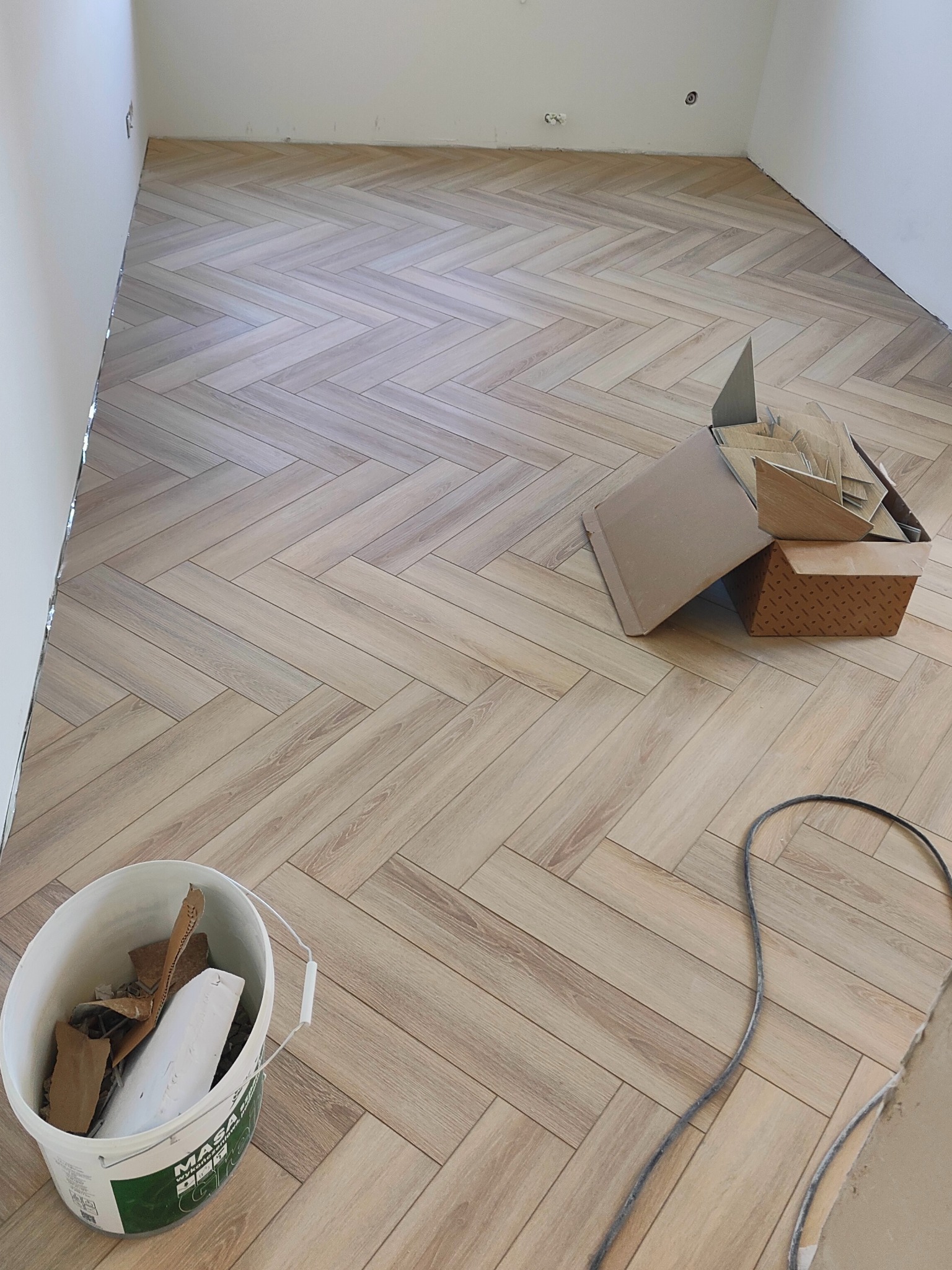 Ułożona podłoga z paneli w jodełkę, widoczny biały kubeł z odpadami i kartony z panelami. Jasne wnętrze w trakcie wykańczania.