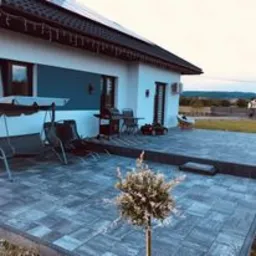 Szara, ułożona kostka brukowa na tarasie przed nowoczesnym domem z białymi ścianami i ciemnym dachem. Widoczne meble ogrodowe, grill i małe drzewko ozdobne. W tle krajobraz wiejski.