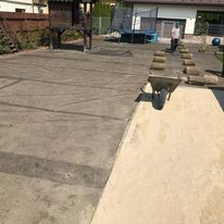 Przygotowanie terenu pod trawnik z użyciem ziemi i rolek trawy, widoczna taczka z piaskiem, w tle dom i trampolina.