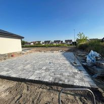 Świeżo ułożona, szara kostka brukowa na podwórku, widoczne obrzeża i przygotowane podłoże, w tle dom jednorodzinny i błękitne niebo.