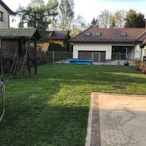 Widok ogrodu z perspektywy tarasu z kostki brukowej, na trawniku domek dla dzieci ze schodkami, w tle dom jednorodzinny z garażem.