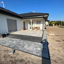 Nowoczesny taras z szarej kostki brukowej ułożony przy białym domu z czarnym dachem. Widoczny fragment ogrodu z przygotowanym podłożem. Cień fotografa na pierwszym planie.