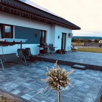 Szara, ułożona kostka brukowa na tarasie przed nowoczesnym domem z białymi ścianami i ciemnym dachem. Widoczne meble ogrodowe, grill i małe drzewko ozdobne. W tle krajobraz wiejski.