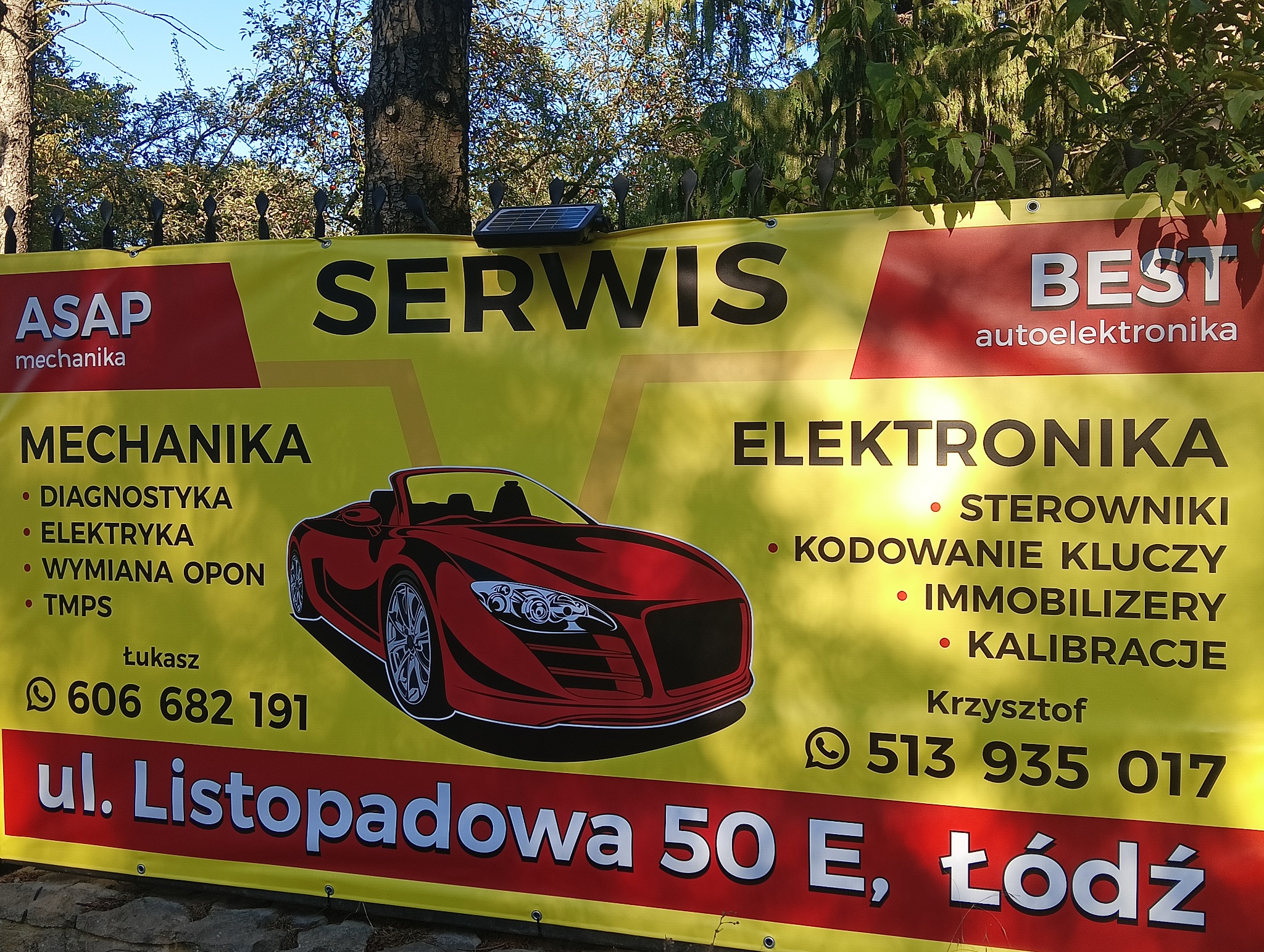 Żółty baner reklamowy serwisu samochodowego ASAP z ofertą usług mechanicznych i elektroniki samochodowej, z ilustracją czerwonego sportowego samochodu i danymi kontaktowymi, zamocowany na zewnątrz...