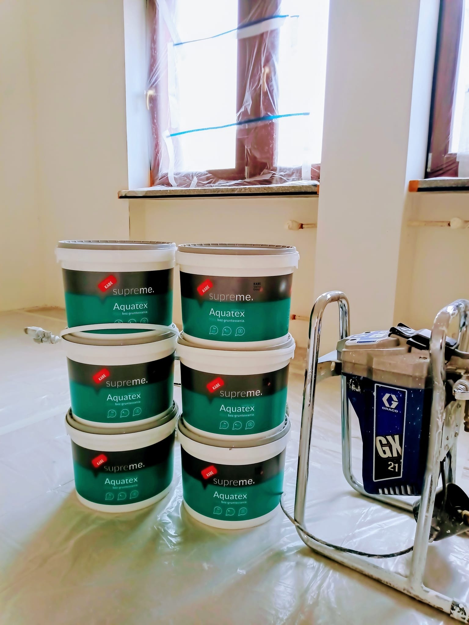 Stos sześciu puszek z zieloną farbą Kabe Supreme Aquatex obok agregatu malarskiego Graco GX21 na folii ochronnej w pomieszczeniu przygotowanym do malowania, okna zabezpieczone folią.