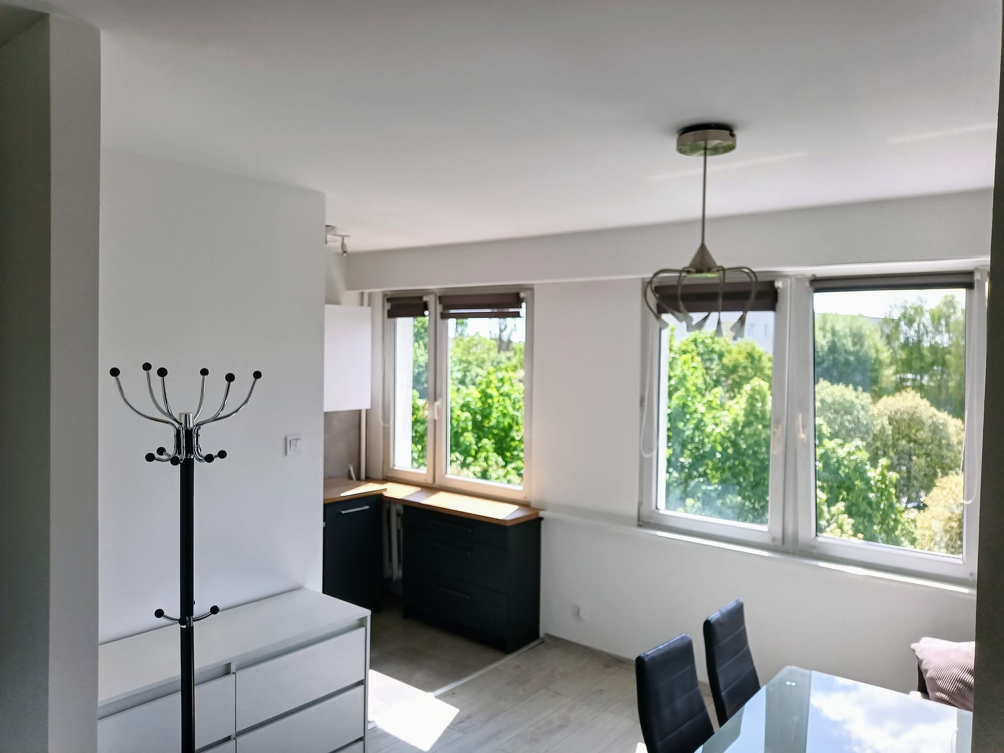 Jasne, odnowione wnętrze mieszkania z widokiem na zieleń za oknem; minimalistyczny design, białe ściany, ciemne meble kuchenne i chromowany wieszak na ubrania przy wejściu.