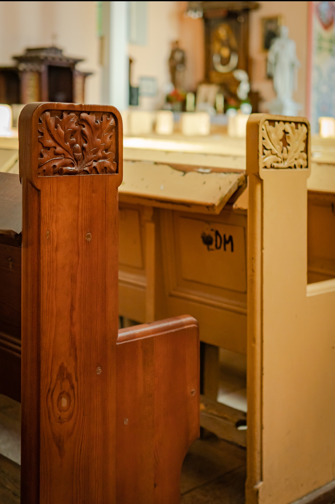 Drewniane ławki kościelne z rzeźbionymi motywami roślinnymi, widoczne ślady użytkowania i napis 'DM' na jednej z ławek.