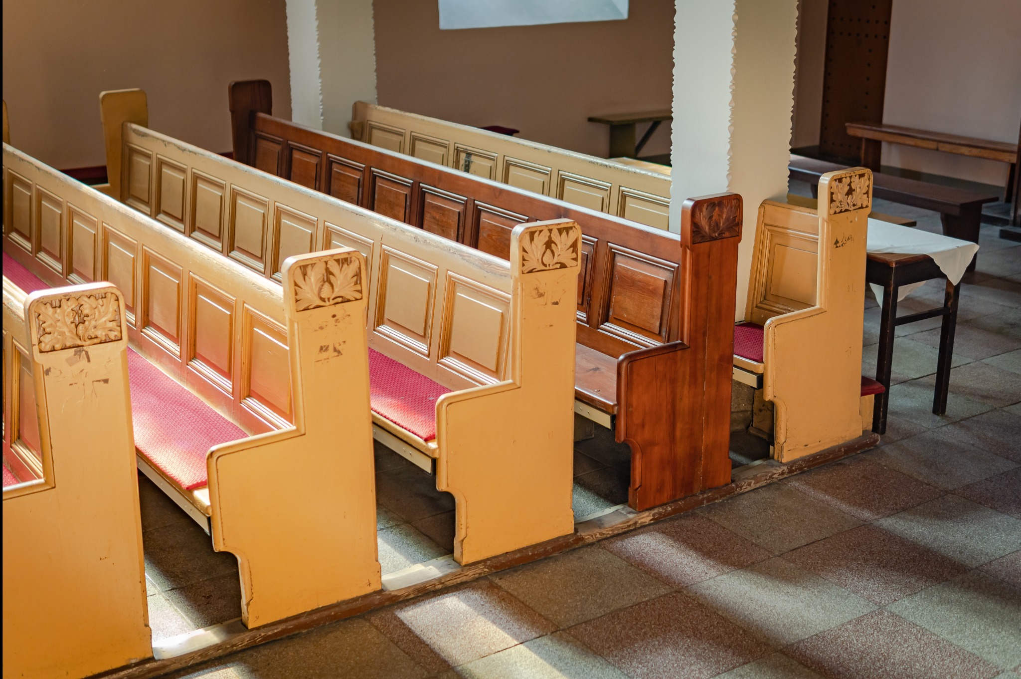 Rząd drewnianych ławek kościelnych z czerwonymi siedziskami, z widocznymi śladami użytkowania i różnymi odcieniami drewna, oświetlonych promieniami słońca padającymi na podłogę.