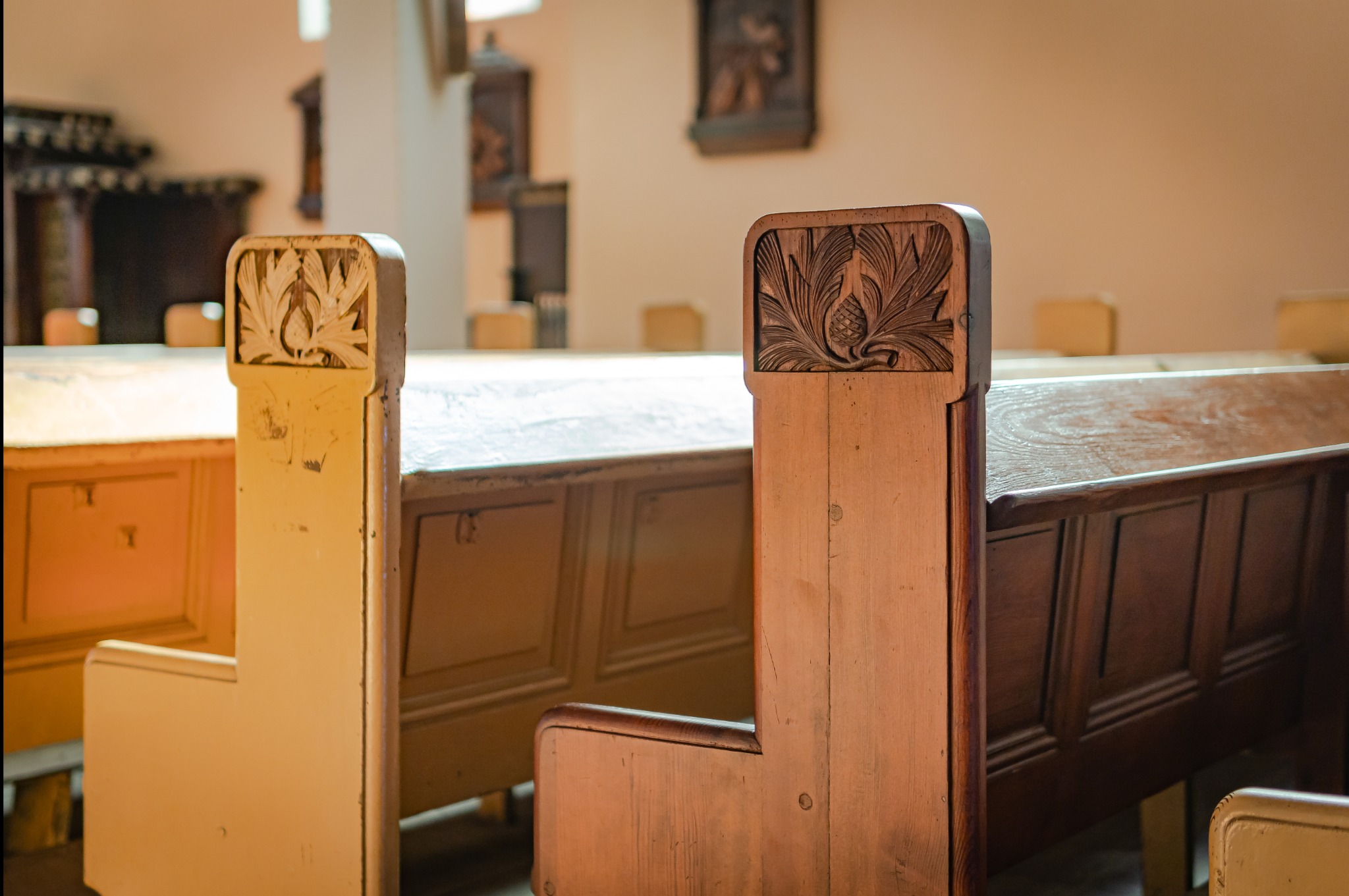 Drewniane ławki w kościele lub innym zabytkowym budynku, z dekoracyjnymi płaskorzeźbami szyszek i liści na oparciach, jedna ławka pomalowana na kremowo, druga w naturalnym kolorze drewna.