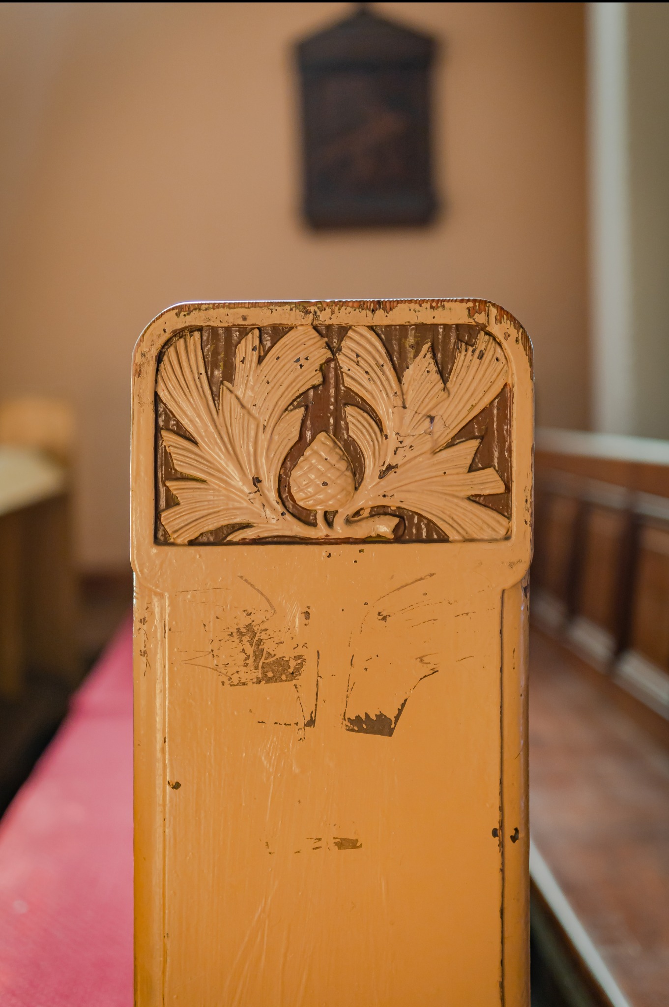 Fragment ławki kościelnej z dekoracyjnym, roślinnym ornamentem i widocznymi śladami użytkowania oraz ubytkami farby, w tle niewyraźny obraz